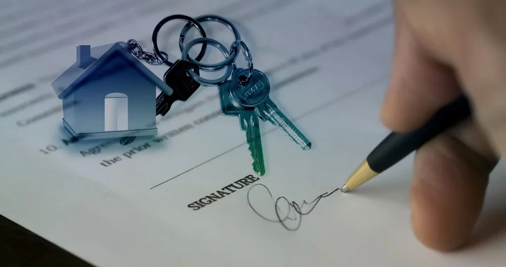 Close-up of a person signing a real estate sale agreement with house keys and keychain – property documentation and legal process by Shikhar Realty
